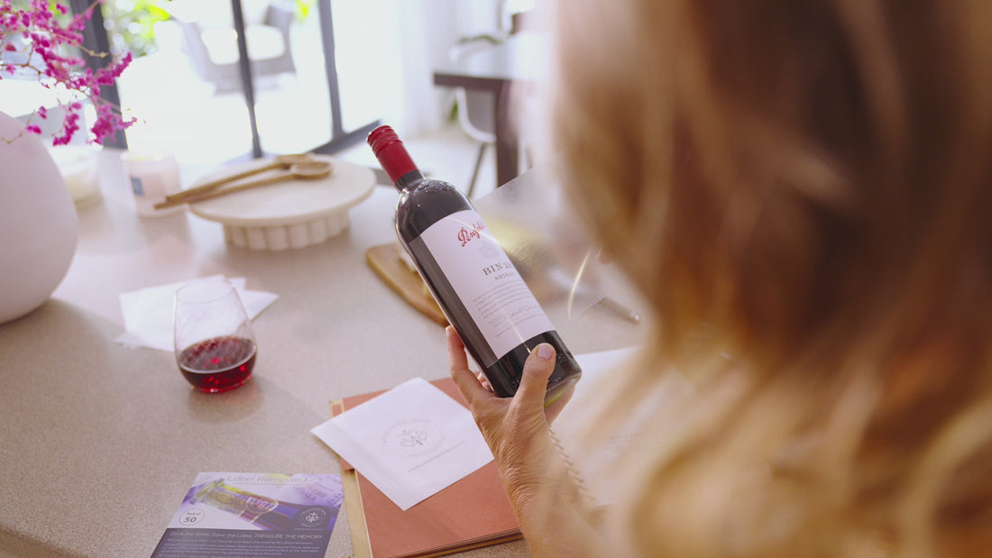 Video showing woman removing a wine bottle label from bottle of Penfolds Bin 28 Shiraz - Woman peeling the backing paper from wine label remover sheet, placing wine label remover sheet over the wine label surface, rubbing the wine label surface, then peeling the wine label remover back from bottle while it removes and laminates the wine label, now ready to place into a wine journal, wine diary, wine card or wine menu